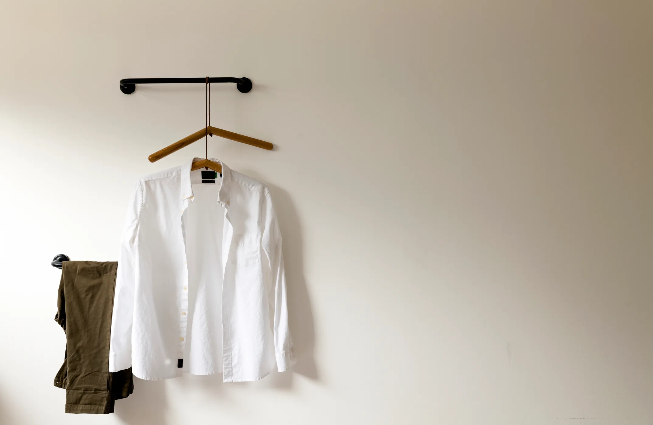 A clean white oxford button-down shirt on a wooden hanger and olive trousers folded on a wall hook against a soft off-white wall — the fashion-editorial endpoint of NYC apparel imports flowing through Port Newark and Elizabeth Marine Terminal from Vietnam, China, India, Indonesia, and Bangladesh.