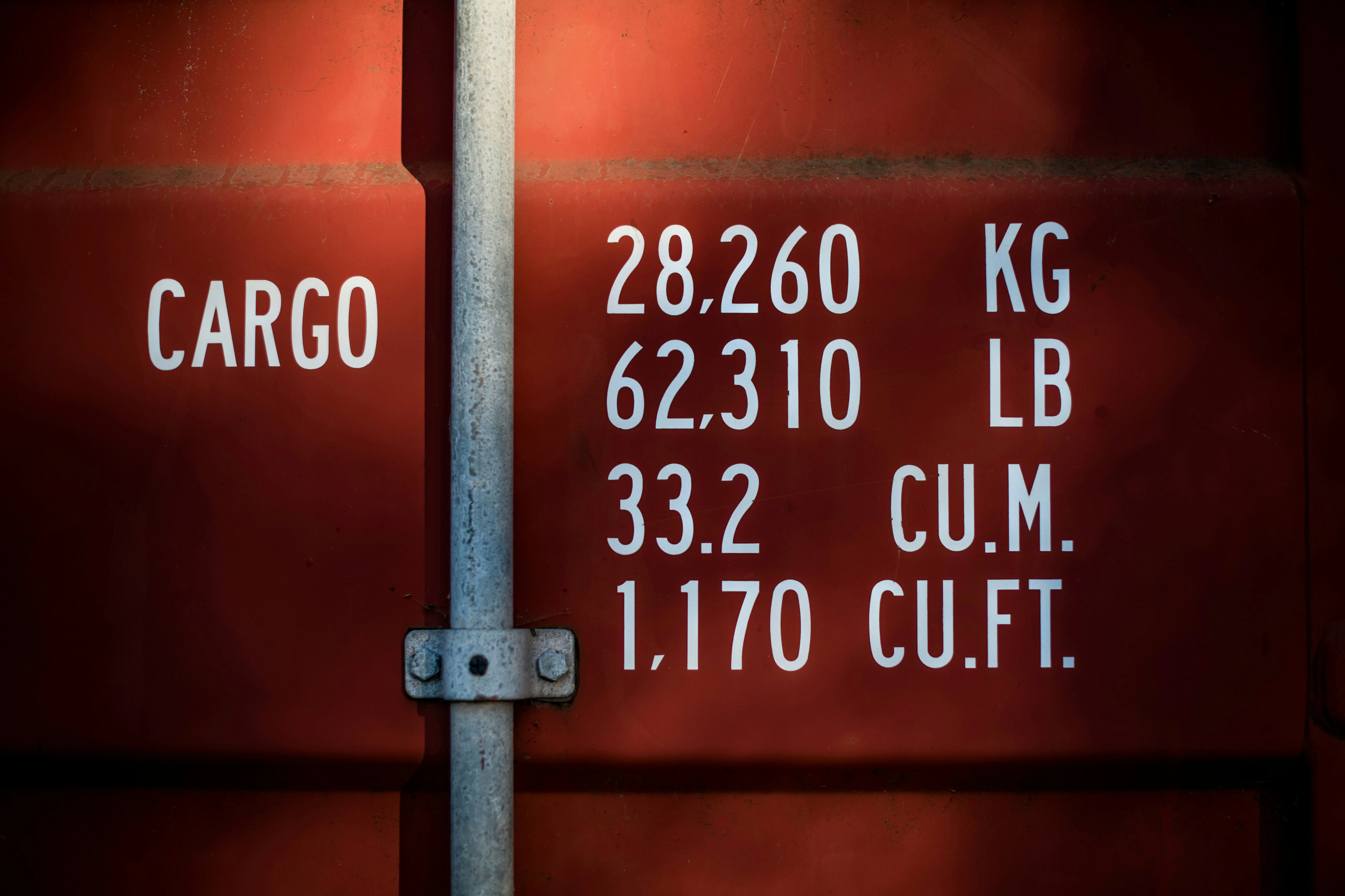 Close-up of a shipping container's stenciled cargo specifications — weight in kilograms and pounds, volume in cubic meters and cubic feet — illuminated by warm side-light. The numerical baseline of every freight engagement.