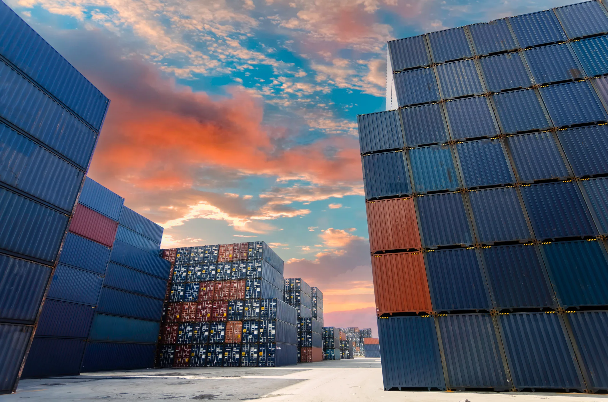 A container yard at sunset — stacked shipping containers in cool blue tones framing a center alley with a warm orange sky, real working harbor infrastructure