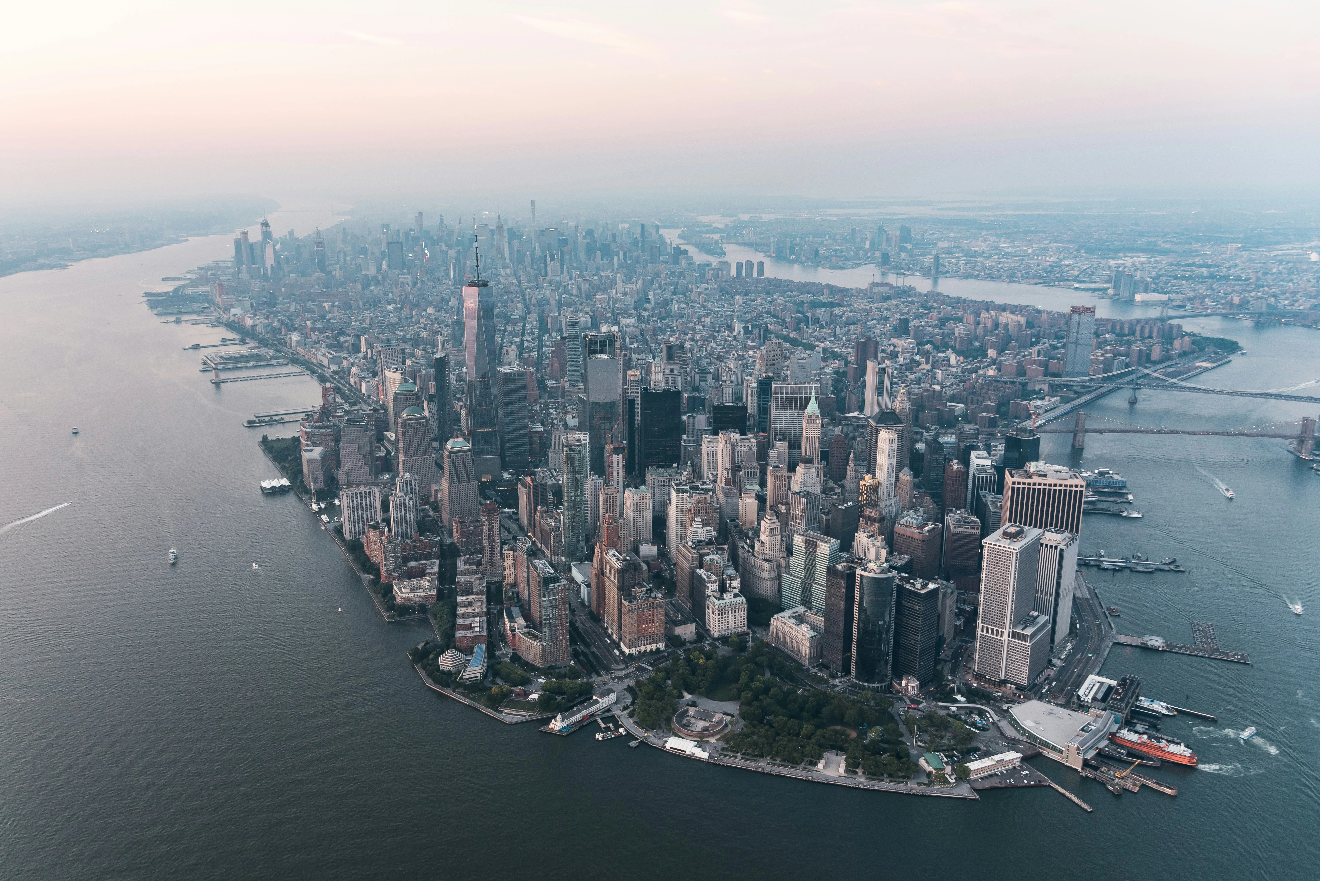 Aerial view of Lower Manhattan at golden hour — One World Trade Center centered in the financial district skyline, the Brooklyn Bridge crossing the East River to the right, the Hudson River and New York Harbor framing the island, with the wider Manhattan grid stretching uptown into a hazy peach-and-grey horizon. The city seen from above; the freight that runs it reported from the floor.