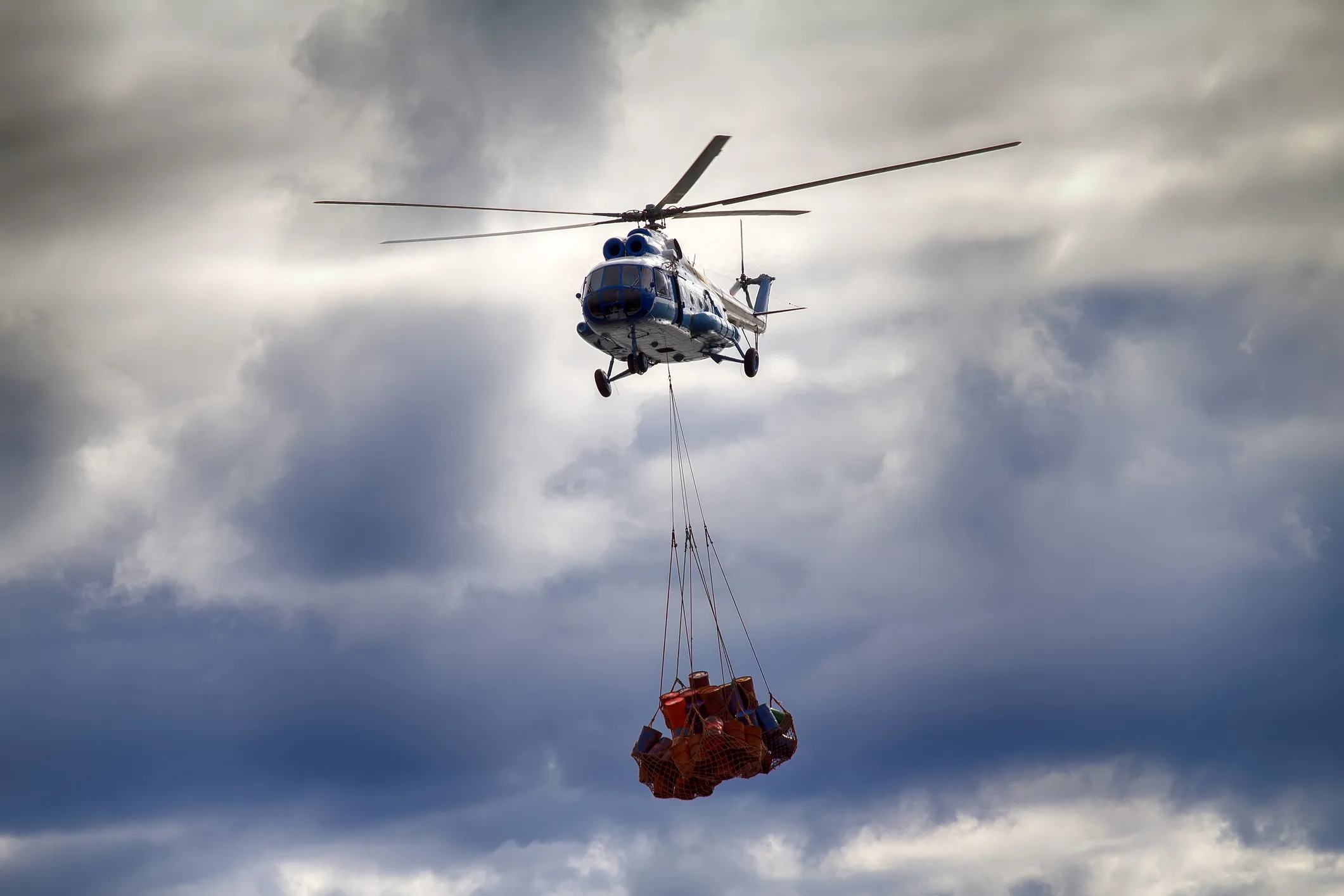 Cargo helicopter in flight carrying a sling-loaded payload — red barrels secured in heavy cargo netting suspended below the airframe — against dramatic cumulus cloud cover. Specialized chartered air lift, one mode in NewYork3PL's international freight forwarding capability set that also includes ocean freight, scheduled air cargo, customs brokerage, and vessel chartering.