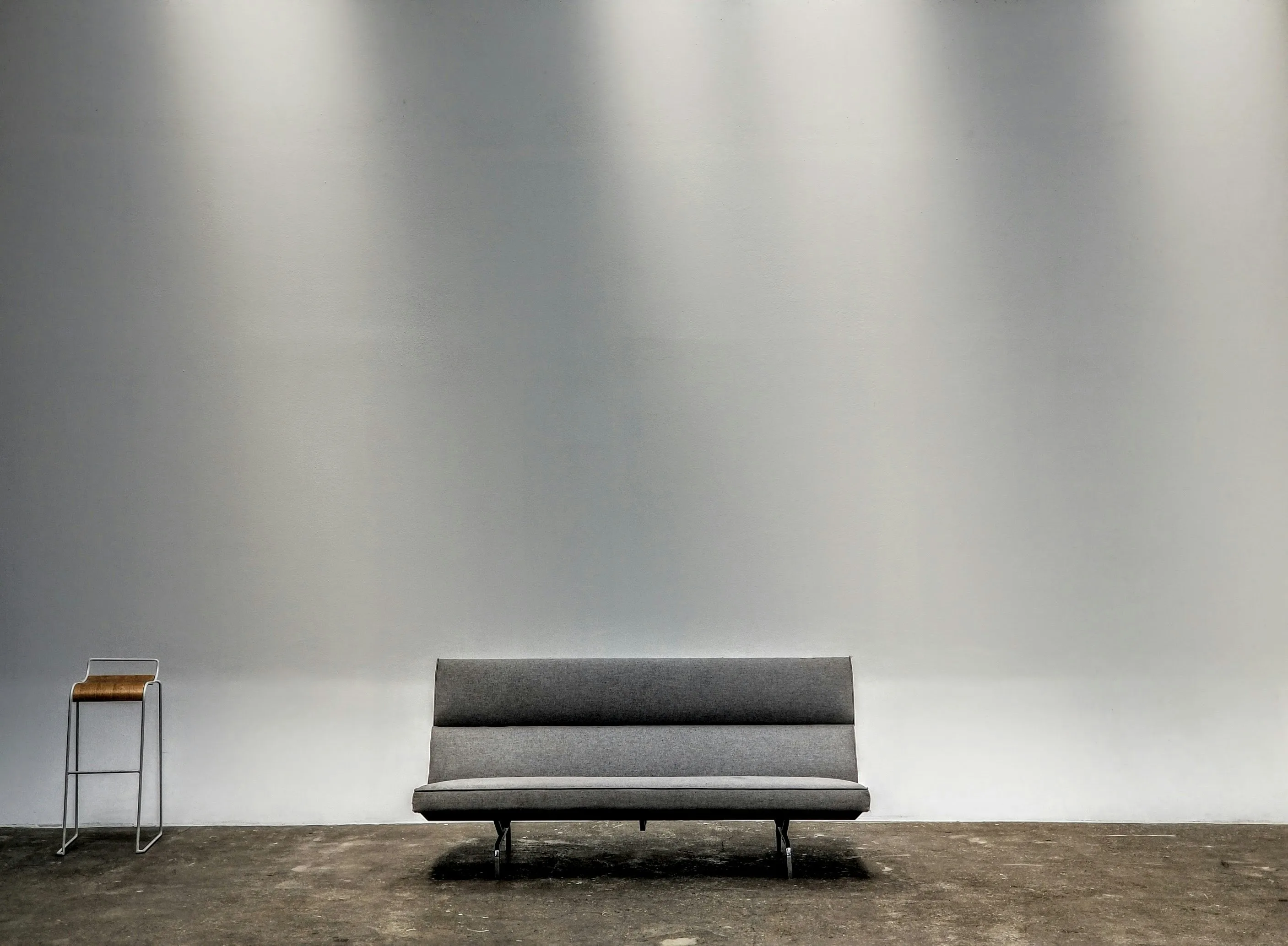 Minimalist gray modern sofa and slim barstool against a soft gray gallery wall on a concrete floor with directional overhead lighting. The high-end residential and design-district endpoint of NYC furniture import logistics — where high-cube containers from Vietnam, China, Indonesia, India, and Thailand land after a multi-step delivery chain.