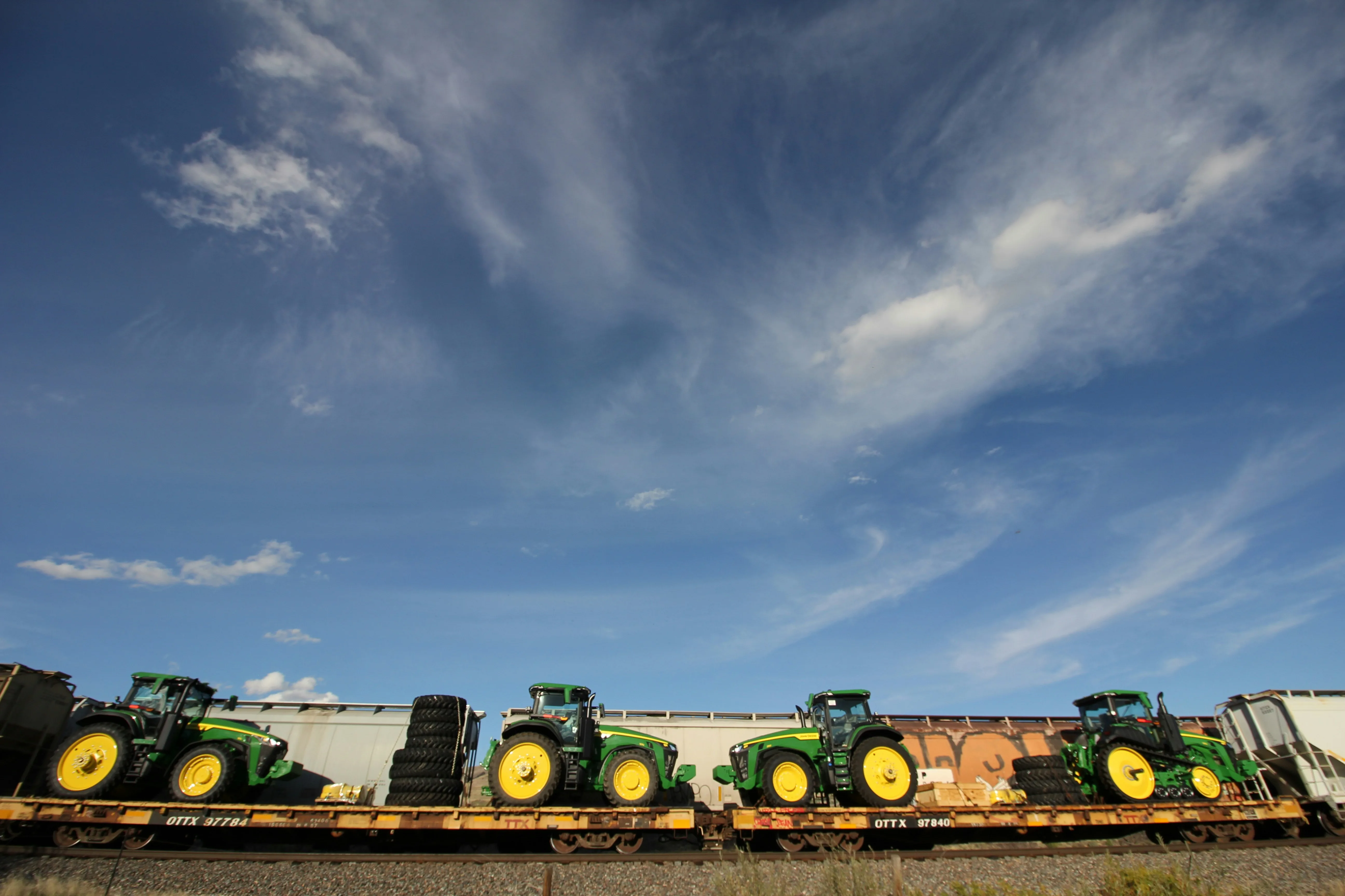 A row of green agricultural tractors loaded on flat-rack railcars under dramatic blue sky with cirrus cloud cover, with rail ballast in the foreground. Heavy machinery and capital equipment imports flowing through Port Newark Container Terminal and onward by rail to Northeast manufacturing customers in NJ, PA, and the broader Mid-Atlantic.