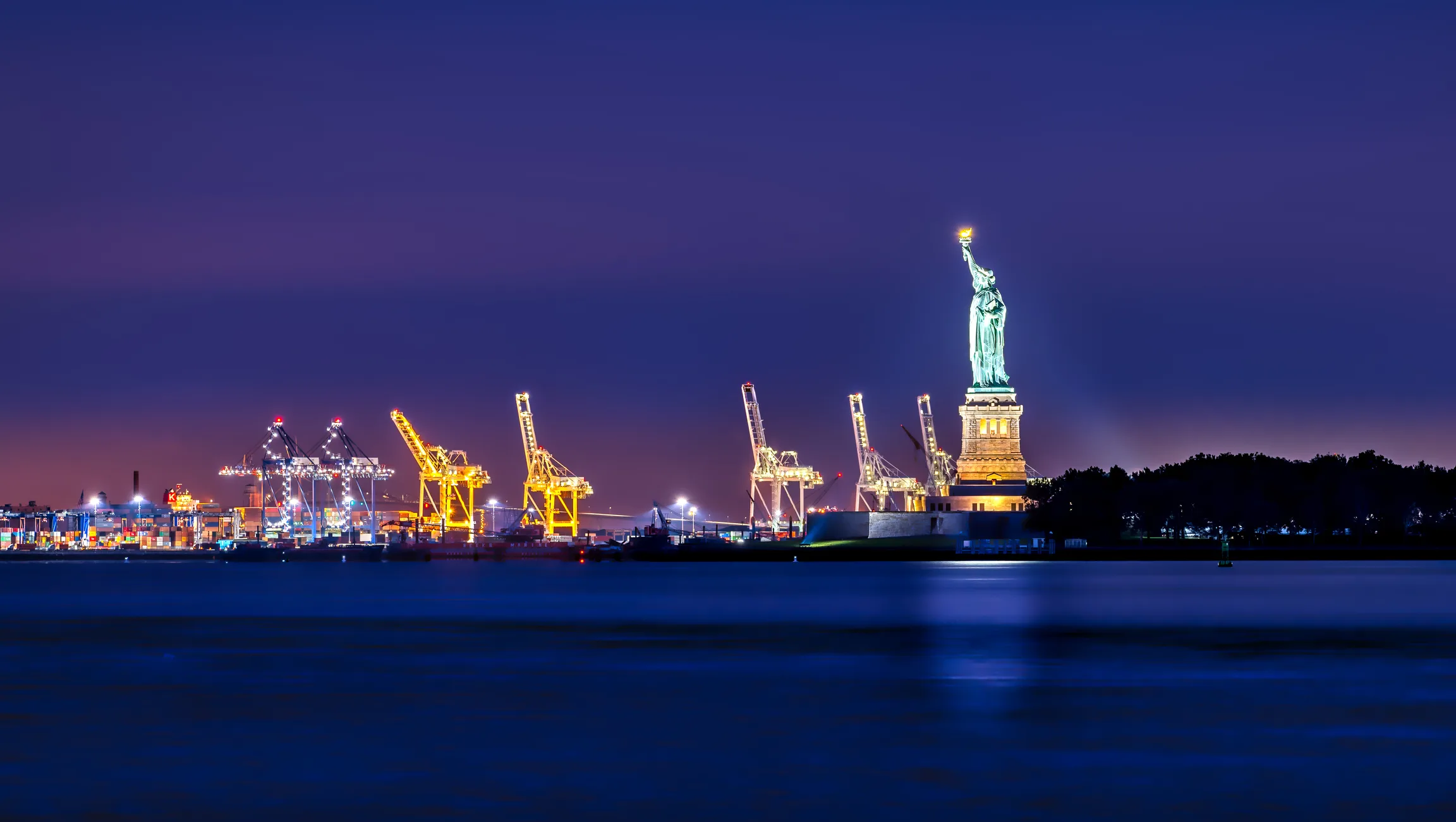 The Port of New York at dusk: illuminated container terminal cranes alongside the Statue of Liberty under a deep purple sky, calm harbor water in the foreground