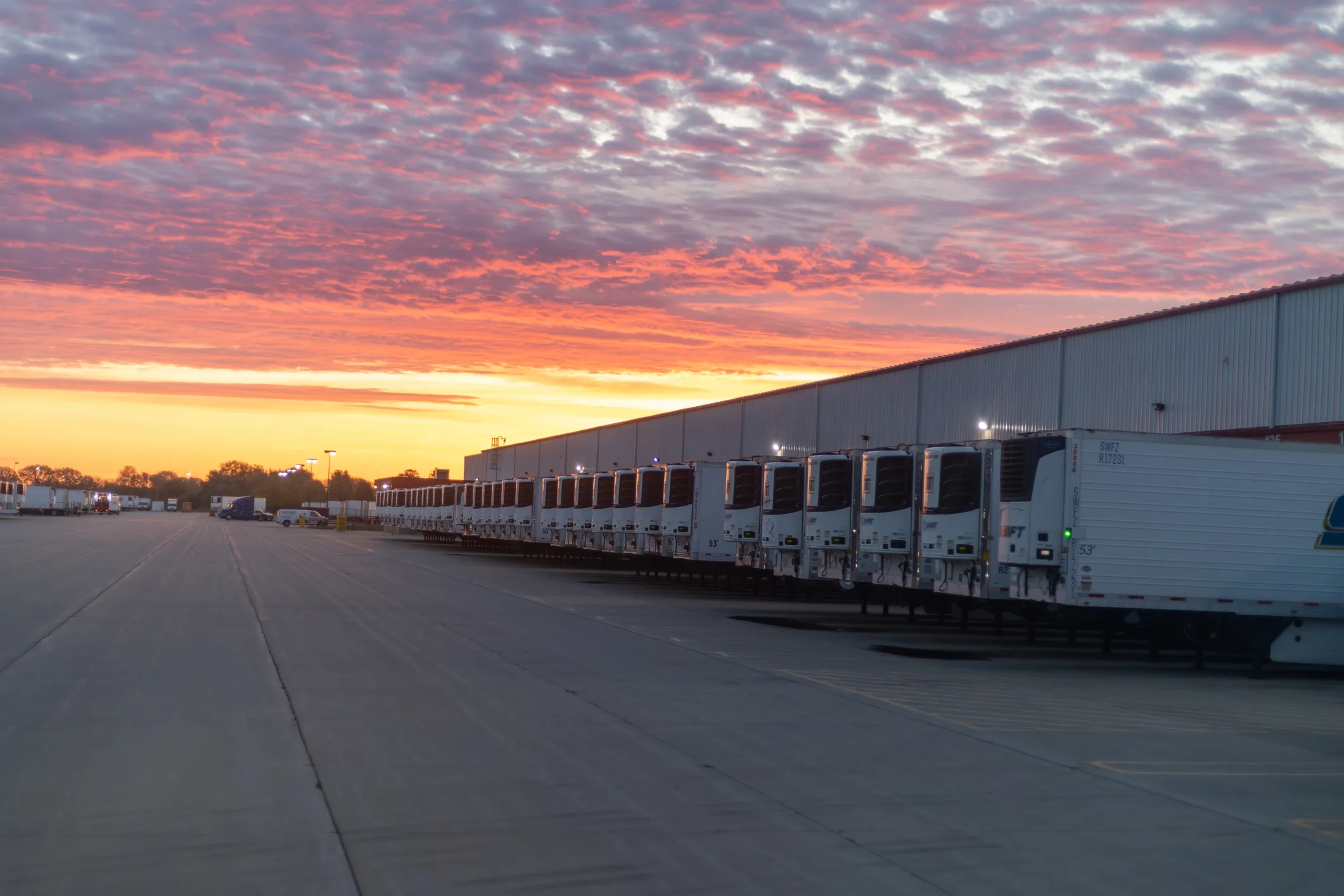 A long row of refrigerated truck trailers (reefer trailers) backed up to a distribution dock at sunrise, with dramatic pink and orange cloud cover overhead and concrete apron in the foreground. The continuous-temperature reefer corridor that operates between PANYNJ and NYC distribution endpoints — pharmaceutical cold-chain, biotech, fresh produce, frozen seafood, dairy, wine, and spirits.
