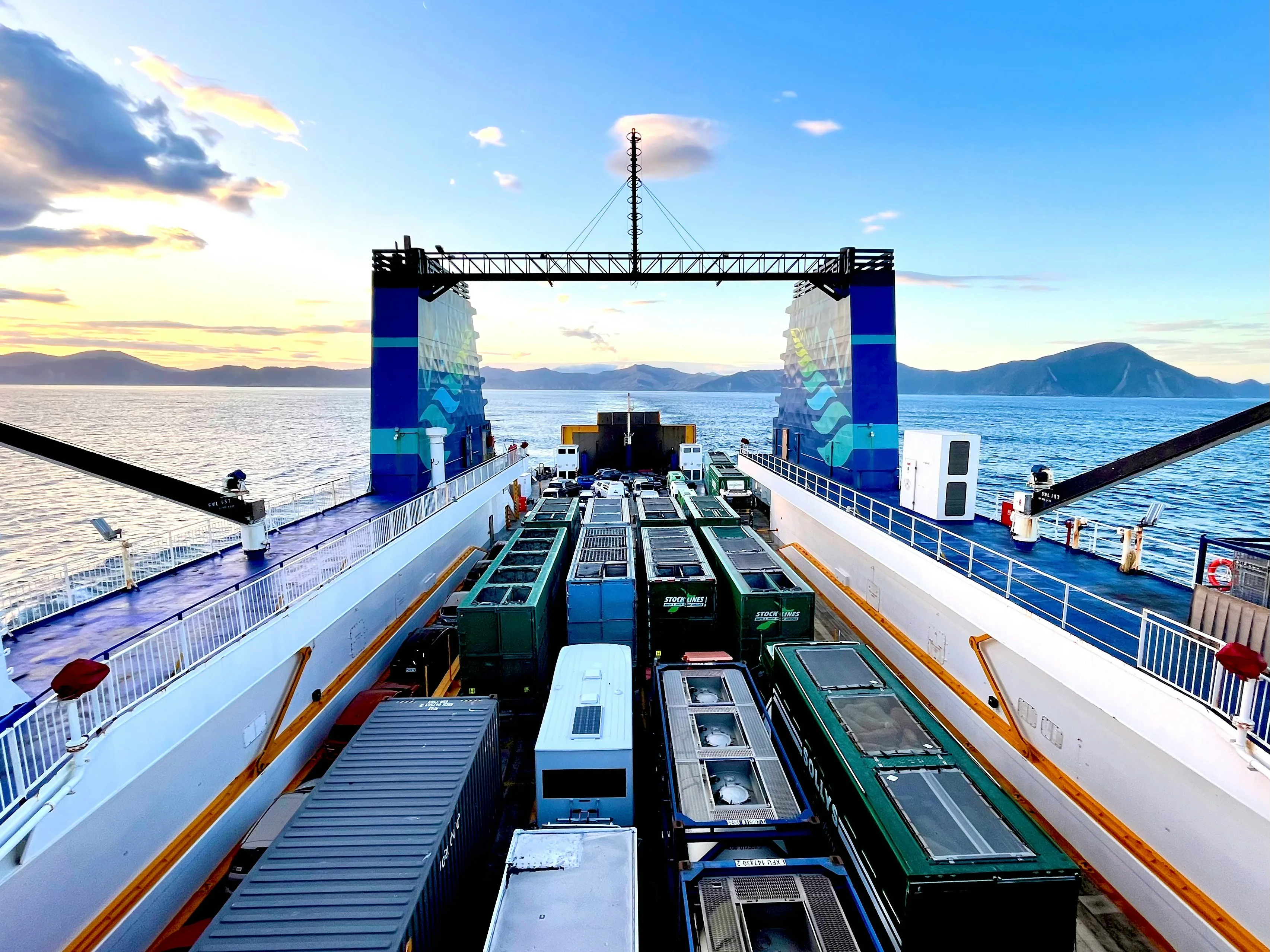 Aerial view from a ro-ro vessel deck looking down at multiple cargo trailers and containers loaded across the ship's deck under golden-hour sky, with coastline and water flanking the vessel. Multi-cargo freight infrastructure typical of the Port of New York and New Jersey throughput across all seven cargo-class sectors NewYork3PL routes.
