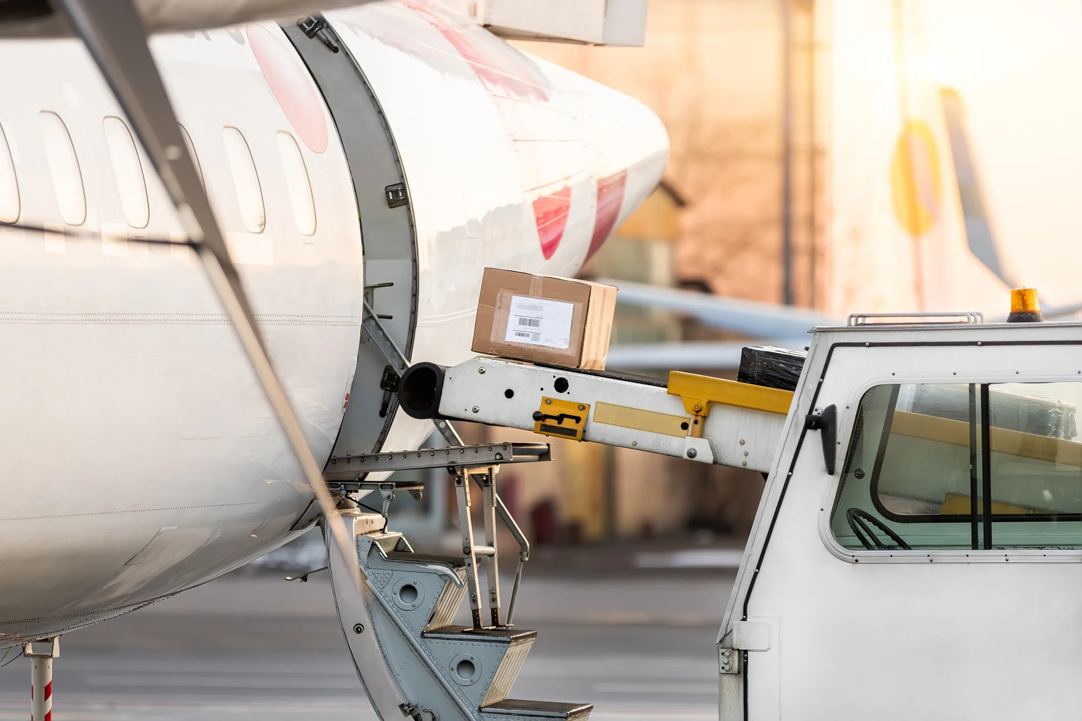 Aircraft fuselage with a parcel conveyor belt mid-load — sealed cardboard boxes traveling into the cargo hold, ground-service tug vehicle in the foreground, golden-hour light on the apron. Specialized cargo handling at an air-cargo terminal — the operational tier where the cargo class itself dictates the protocol.