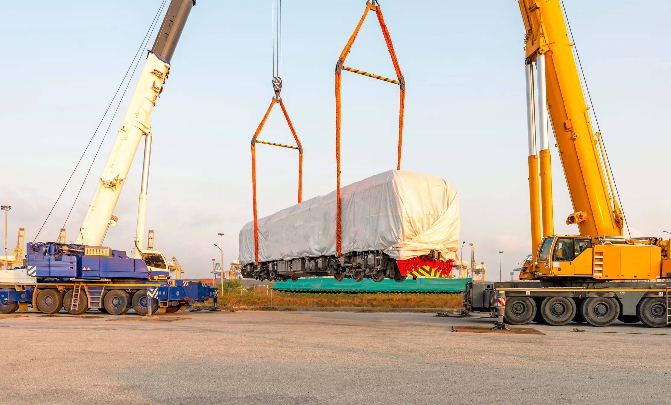 Two industrial cranes lifting a wrapped and tarped piece of project cargo with red and orange rigging slings, on a concrete port apron with port infrastructure visible in the background. The bespoke heavy-lift handling that specialty and project cargo requires — beyond standard containerized shipping.