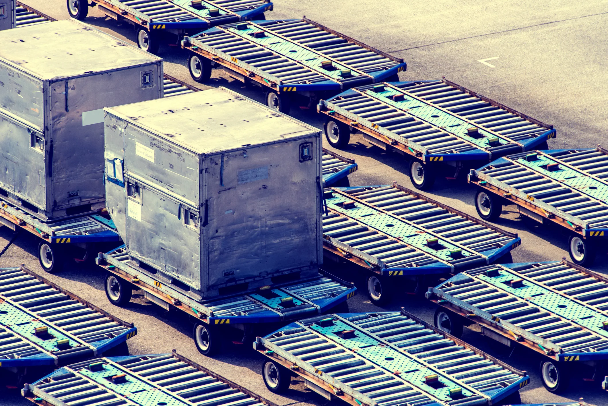 Overhead view of an airport ground-handling apron — rows of aluminum ULD cargo containers and conveyor dollies arranged in repeating geometric pattern. Industrial multi-mode freight infrastructure typical of the JFK and EWR air cargo terminals NewYork3PL operates through, alongside truck, rail, and ocean carriers.