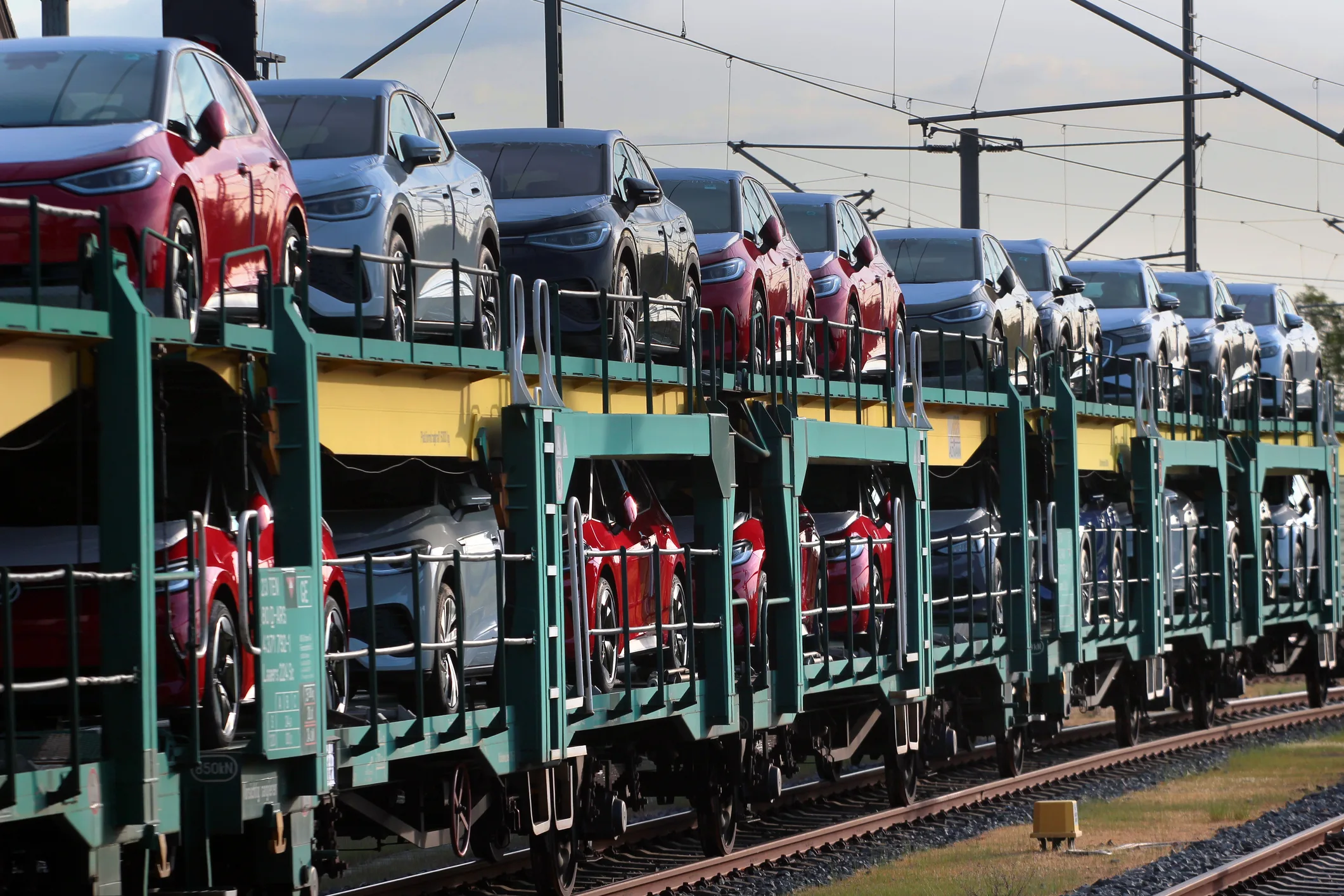 A row of new passenger vehicles loaded on multi-tier auto-rack railcars at a rail yard, with overhead catenary wires above and gravel ballast in the foreground. Vehicle imports flowing through the Port Authority Auto Marine Terminal and onward by rail and auto-haul carrier to dealer networks across the Northeast.