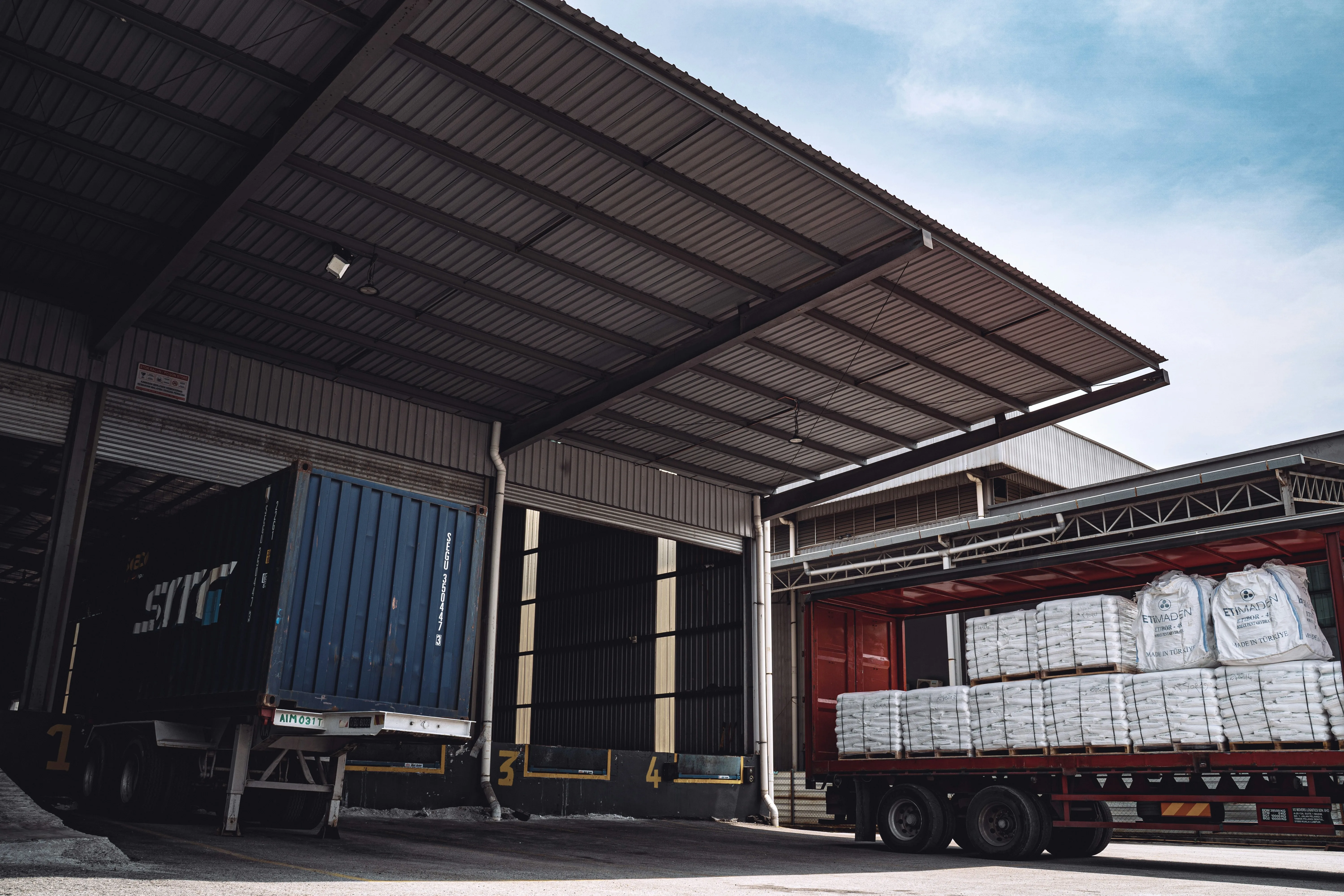 Warehouse loading dock complex with multiple numbered bays — bay one with a blue intermodal container in mid-load, bay four with a flatbed trailer carrying palletized white-bagged cargo — under a daytime sky. Industrial-grade multi-bay infrastructure typical of the multi-borough partner facilities NewYork3PL operates across Brooklyn, Queens, Bronx, Staten Island, Northern New Jersey, and Lehigh Valley.